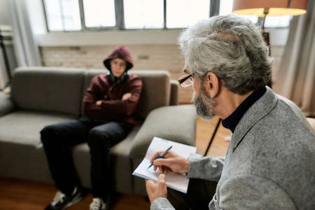 Psychoanalyst taking notes during appointment with uneasy teenage boy