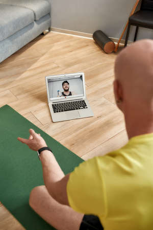 Sport and healthy lifestyle. Male fitness coach sitting on yoga mat at home, explaining workout techniques online while having video conference on laptopの写真素材