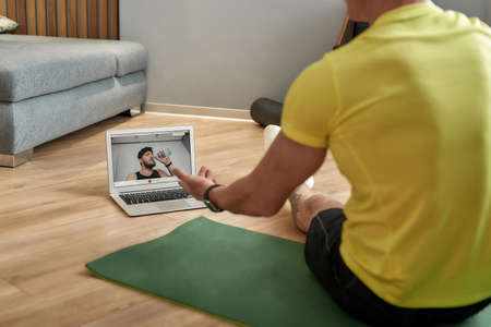 Your personal trainer online. Rear view of a mature fitness coach in sportswear sitting on yoga mat at home and conducting online training, having virtual fitness classの写真素材
