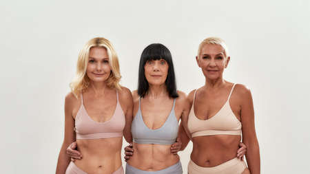 Beautiful senior women. Three attractive middle aged female friends in underwear embracing each other while posing together on light backgroundの写真素材