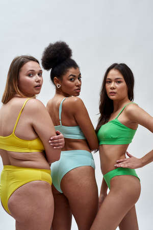 Three multiethnic young women with different body shapes in colorful underwear turning their heads to camera, posing isolated over light backgroundの写真素材