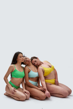 Full length shot of three diverse young women in colorful underwear posing together, leaning on each other, sitting on knees with eyes closed isolated over white backgroundの写真素材