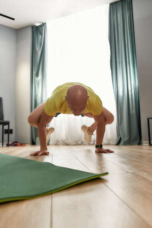 Front view of sportive bald man working out, standing on his arms and legs in the air while training in living room at homeの写真素材