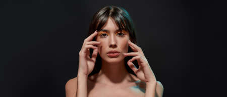 Beauty portrait of young brunette woman with healthy brown hair and professional makeup looking at camera, posing with hands near her face isolated over black backgroundの写真素材