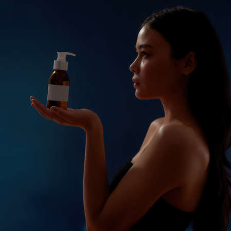 Skin care concept. Young sensual asian woman holding cosmetic product on the palm and looking at it while standing against dark blue backgroundの写真素材