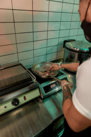 High angle view of male cook wearing protective mask and gloves frying pork or beef meat in wok pan...
