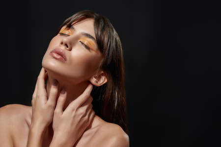 Beauty portrait of young brunette woman with perfect skin and professional orange makeup posing with eyes closed and hands on her neck isolated over black backgroundの写真素材