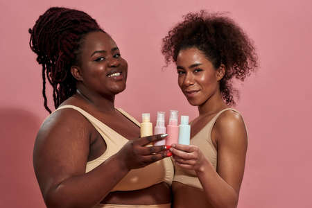 Two young afro female wearing beige underwear and holding bottles of care productの写真素材