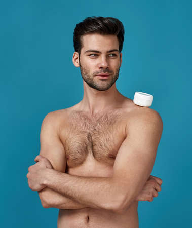 Portrait of sexy brunette guy posing with white jar of effective cream for face on his shoulder, looking aside, standing with arms crossed isolated over blue backgroundの写真素材