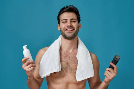 Attractive guy with white towel around his neck smiling at camera, holding a shaving foam and electric shaver, posing isolated over blue backgroundの写真素材