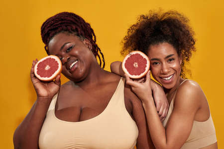 Two excited young african american women in underwear smiling at camera, holding halfs of juicy grapefruit, posing together isolated over orange backgroundの写真素材