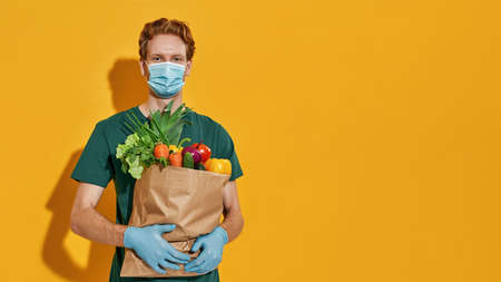 Young courier in mask holding packet with green grocery