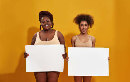 Two cheerful different african american women in underwear laughing at camera, holding blank banner for your text, standing isolated over yellow backgroundの写真素材
