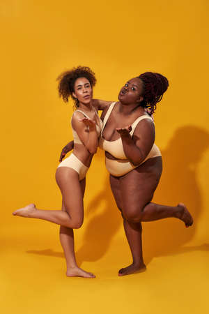 Two cheerful young african american women in underwear with different body size blowing kiss while posing together isolated over yellow backgroundの写真素材
