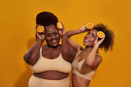 Two playful african american women having fun, holding half of ripe juicy orange, posing together isolated over orange backgroundの写真素材