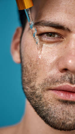 Half face closeup of attractive man with smooth skin looking at camera, applying serum beauty product on his face using glass pipette isolated on blue backgroundの写真素材