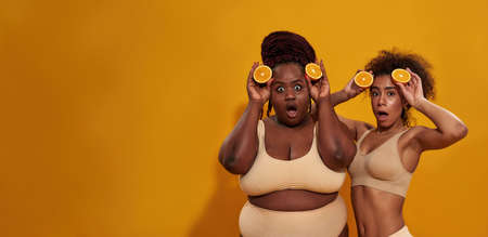 Two funny young african american women looking surprised at camera, holding halves of ripe juicy orange near their faces, posing isolated over orange backgroundの写真素材