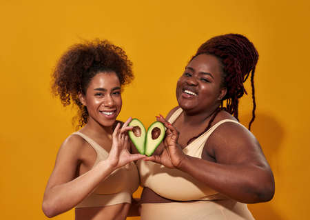 Two african american women in underwear smiling at camera, holding healthy fruit avocado halves as a...