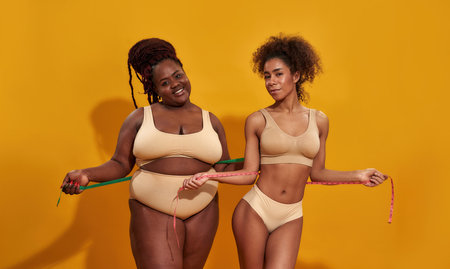Studio shot of cheerful african american women in underwear with different body shape holding metric tape measure, standing isolated over orange backgroundの写真素材
