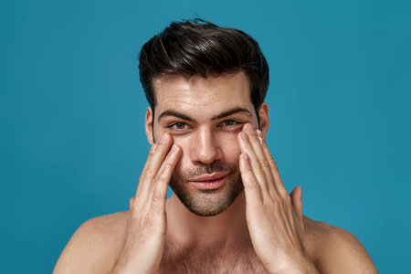 Beauty portrait of handsome man looking at camera with a smile while applying moisturizing cream on his face isolated over blue backgroundの写真素材