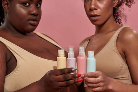 Two young afro female wearing beige tops and showing cosmetics, focus on the bottlesの写真素材