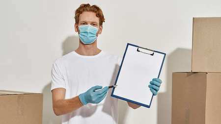 Young redhead courier holds a clipboard and pen, wearing gloves and mask