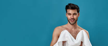 Studio portrait of cheerful brunette guy using a white towel to wipe himself after shaving and taking a morning shower, smiling at camera, posing isolated over blue backgroundの写真素材