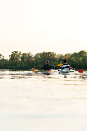 Rear view of two young guys best friends paddling kayak on a lake surrounded by peaceful nature