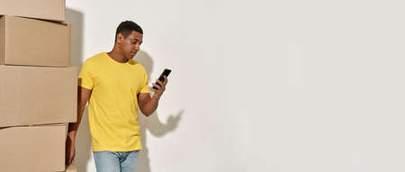 Young male professional mover in casual wear using smartphone, standing near a stack of cardboard...