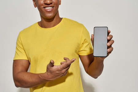 Cropped shot of smiling young man in casual yellow t shirt showing and pointing at smartphone with...
