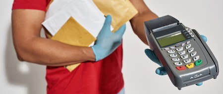 Close up of delivery man wearing protective gloves holding parcels, envelopes and payment terminal...