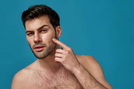 Portrait of shirtless handsome caucasian man checking his facial skin isolated over blue backgroundの写真素材