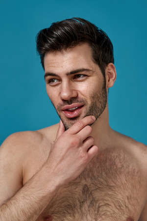 Portrait of handsome naked brunette guy with fresh skin looking aside, touching his chin while posing isolated over blue backgroundの写真素材