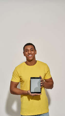 Cheerful young man in casual yellow t shirt smiling at camera, showing digital tablet pc with blank screen while standing isolated over gray backgroundの写真素材