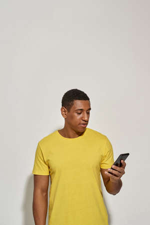 Focused young man in casual yellow t shirt looking busy, holding and using his smartphone while...