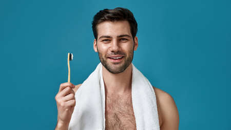 Close up portrait of cheerful guy with towel around his neck smiling at camera, holding toothbrush while posing isolated over blue backgroundの写真素材