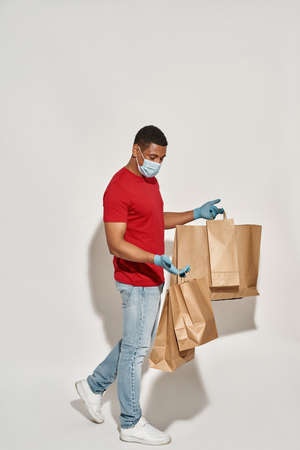 Full length shot of delivery man in protective mask and gloves holding many brown craft paper bags...