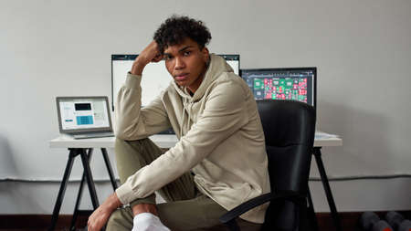 Thoughtful young guy, mixed race trader working from home using laptop and computer, sitting in...