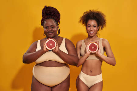 Two young african american women in underwear smiling at camera, holding halfs of juicy grapefruit in front of them, posing isolated over orange backgroundの写真素材