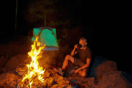 Tired male tourist drinking hot tea or coffee while sitting near campfire outdoors in forest at...