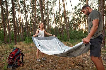 Young couple of tourists looking focused while setting up a tent together in forest