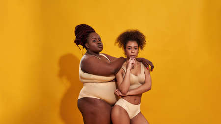 Two serious african american women in beige underwear with different body size standing together,...