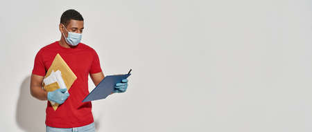 Busy delivery man wearing face mask and protective gloves looking at clipboard, holding parcels and...