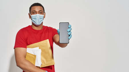 Delivery man wearing face mask and protective gloves looking at camera, holding parcels, letters and...