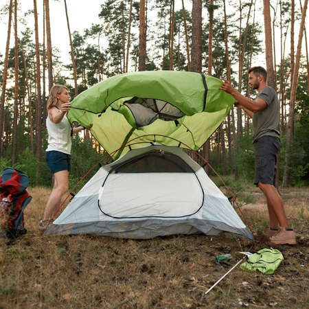 Full length shot of young couple of tourists setting up camping tent in forestの写真素材