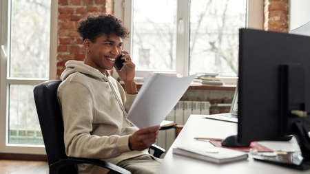 Successful young male trader holding papers, smiling while talking on the phone with a client, sitting at desk, trading from homeの写真素材