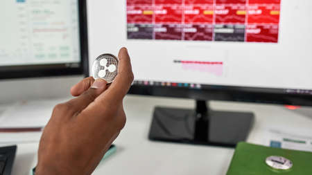 Close up of hand of male trader holding Ripple XRP cryptocurrency token, working on computer while...