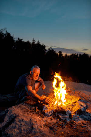 Mature man, tourist looking busy while making a fire, spending time outdoors in forest at twilight
