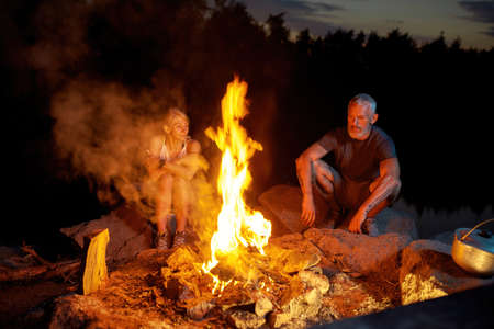A couple of tourists relaxing near campfire while spending time outdoors in forest