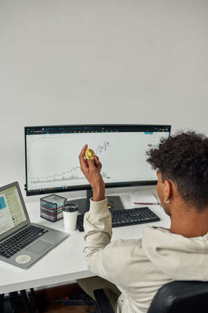 Back view of busy male trader holding Ethereum ETH cryptocurrency coin, studying charts while trading from homeの写真素材